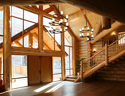 The front of the handcrafted log house is almost entirely windows framed by logs. Large dramatic spiral wrought iron chandeliers hang from the tall ceiling.